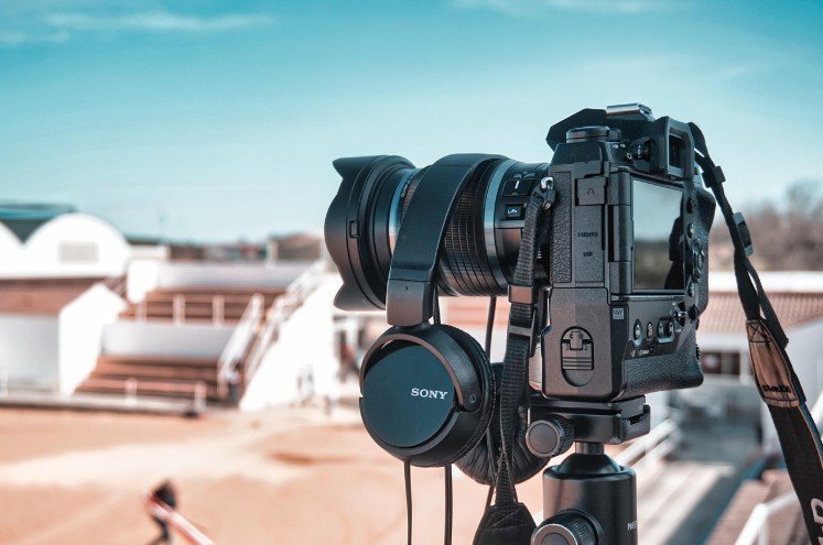 Un appareil photo Sony noir sur un trépied auquel sont suspendus des écouteurs Sony, installé en plein air près d'un terrain de sport avec des sièges vides en arrière-plan.
