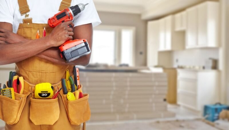 Une personne portant une ceinture à outils tient une perceuse sans fil à l'intérieur d'une pièce en cours de rénovation, avec des matériaux de construction et des armoires inachevées à l'arrière-plan.