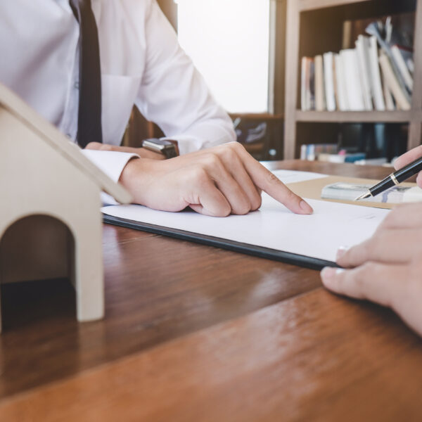 Deux personnes examinent un document et en discutent à un bureau où se trouve une petite maquette de maison en bois, suggérant une réunion sur l'immobilier ou un contrat de logement.