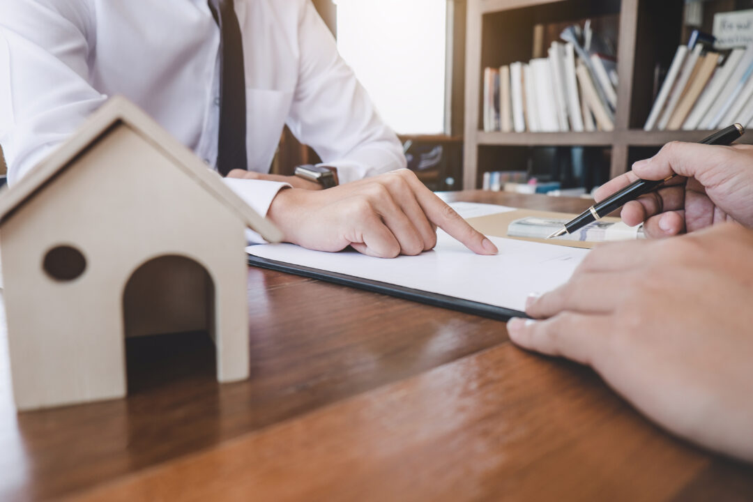 Deux personnes examinent un document et en discutent à un bureau où se trouve une petite maquette de maison en bois, suggérant une réunion sur l'immobilier ou un contrat de logement.