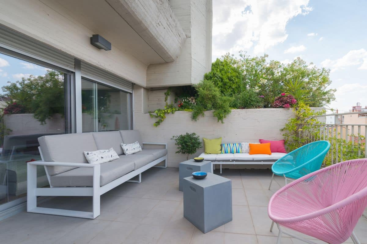 Terrasse moderne avec canapé gris, coussins colorés, deux chaises roses et bleues et une table grise. Des plantes luxuriantes ornent les murs et des portes vitrées s'ouvrent sur l'intérieur. Vivez une ambiance de terrasse en 2025 sous un ciel clair et lumineux.