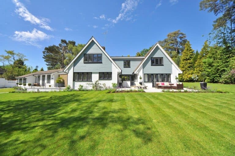 Une grande maison de banlieue avec deux toits pointus, un extérieur blanc et bleu clair, entourée d'une pelouse verte bien entretenue et d'arbres sous un ciel bleu clair.