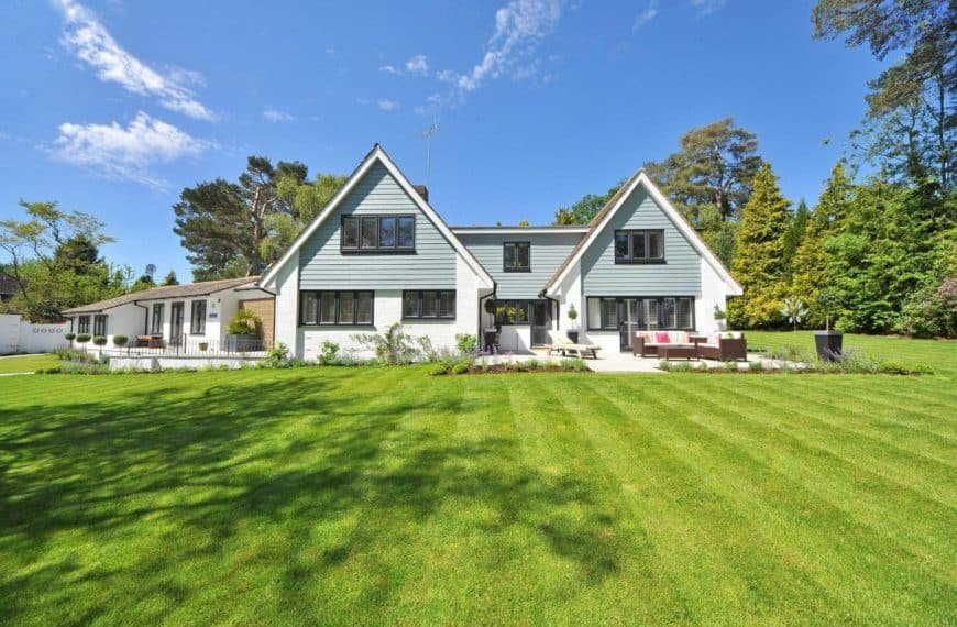 Une grande maison de banlieue avec deux toits pointus, un extérieur blanc et bleu clair, entourée d'une pelouse verte bien entretenue et d'arbres sous un ciel bleu clair.