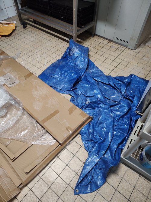 Une bâche bleue est étalée et partiellement froissée sur un sol carrelé, à côté de cartons aplatis.