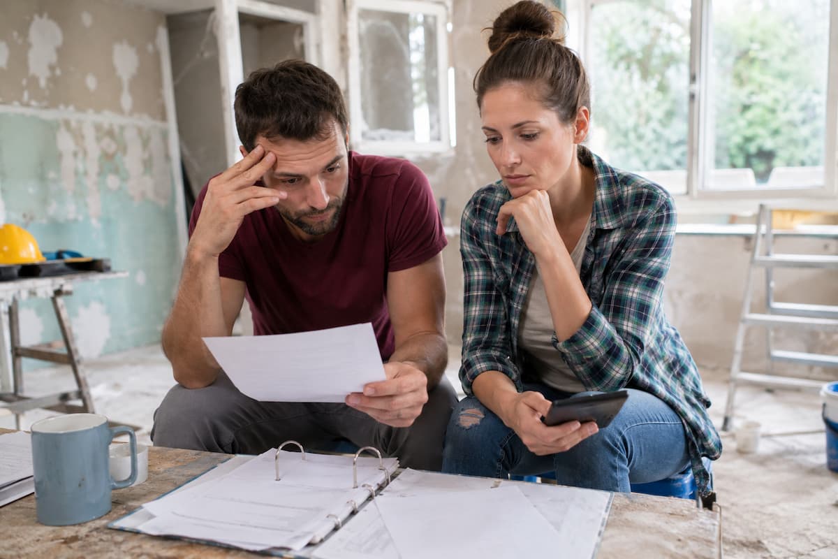 Un homme et une femme sont assis dans une pièce partiellement rénovée ; ils consultent des documents et un classeur, tandis que la femme tient un smartphone à la main et que l'on aperçoit des matériaux de construction à l'arrière-plan.