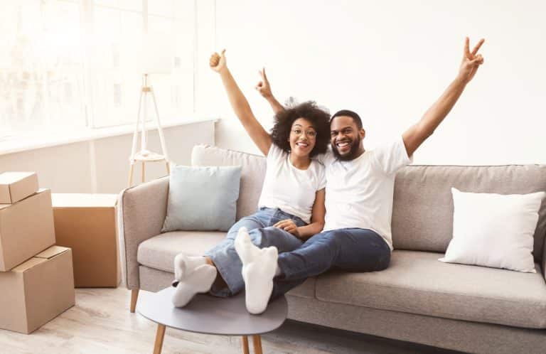 Un couple souriant, assis sur un canapé, les bras levés pour célébrer. Ils sont entourés de cartons de déménagement dans une pièce bien éclairée.