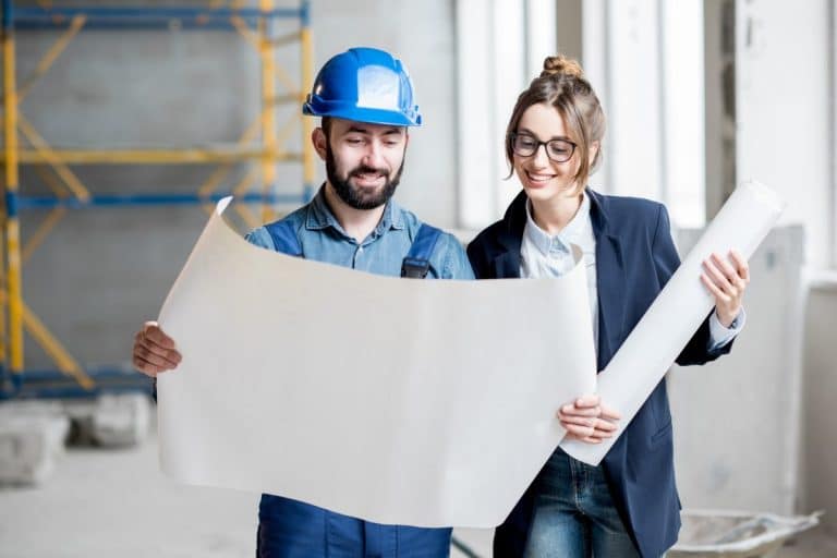 Un homme et une femme examinent des plans sur un chantier. L'homme porte un casque bleu et une salopette, tandis que la femme porte des lunettes et un blazer. Un échafaudage est visible en arrière plan.