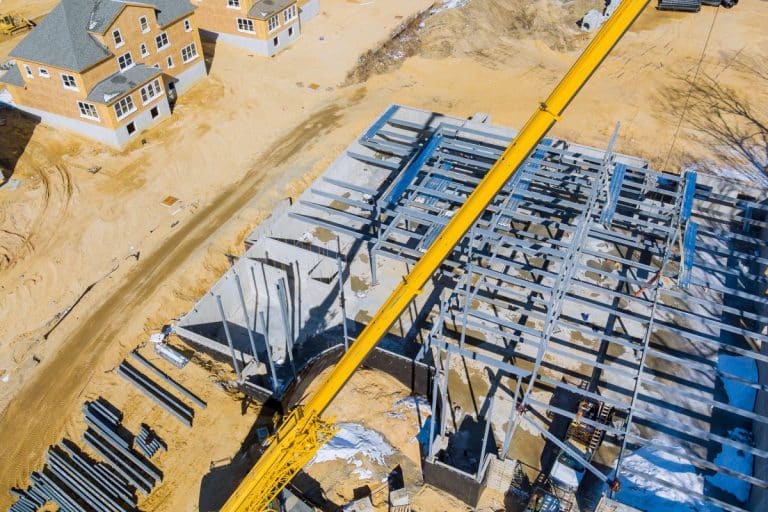 Chantier de construction avec une charpente métallique assemblée par une grue. A proximité se trouvent des maisons partiellement construites sur un sol sablonneux.