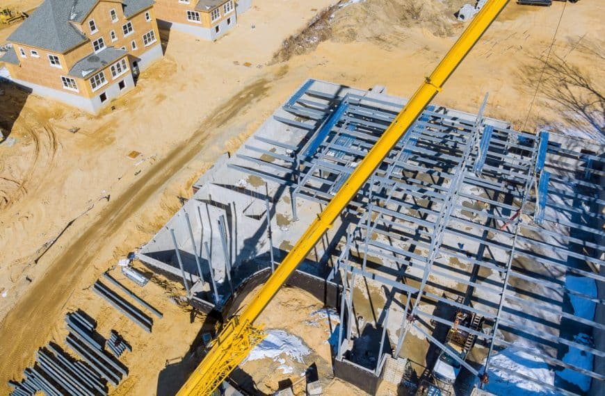 Chantier de construction avec une charpente métallique assemblée par une grue. A proximité se trouvent des maisons partiellement construites sur un sol sablonneux.