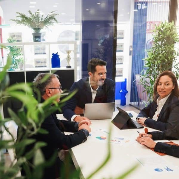 Cinq personnes en tenue professionnelle en réunion autour d'une table avec des ordinateurs portables et des documents dans un espace de bureau lumineux avec des plantes.