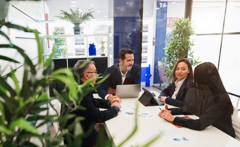 Cinq personnes en tenue professionnelle en réunion autour d'une table avec des ordinateurs portables et des documents dans un espace de bureau lumineux avec des plantes.
