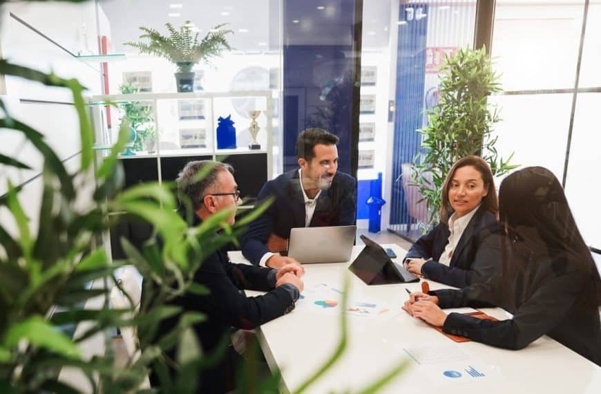 Cinq personnes en tenue professionnelle en réunion autour d'une table avec des ordinateurs portables et des documents dans un espace de bureau lumineux avec des plantes.
