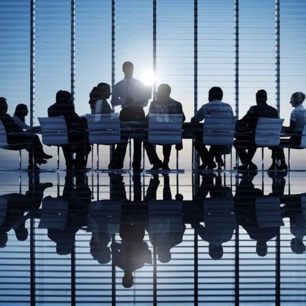 Silhouette d'un groupe de personnes assises autour d'une table de conférence dans un bureau moderne avec de grandes fenêtres et une vue sur la ville, une personne debout et parlant.