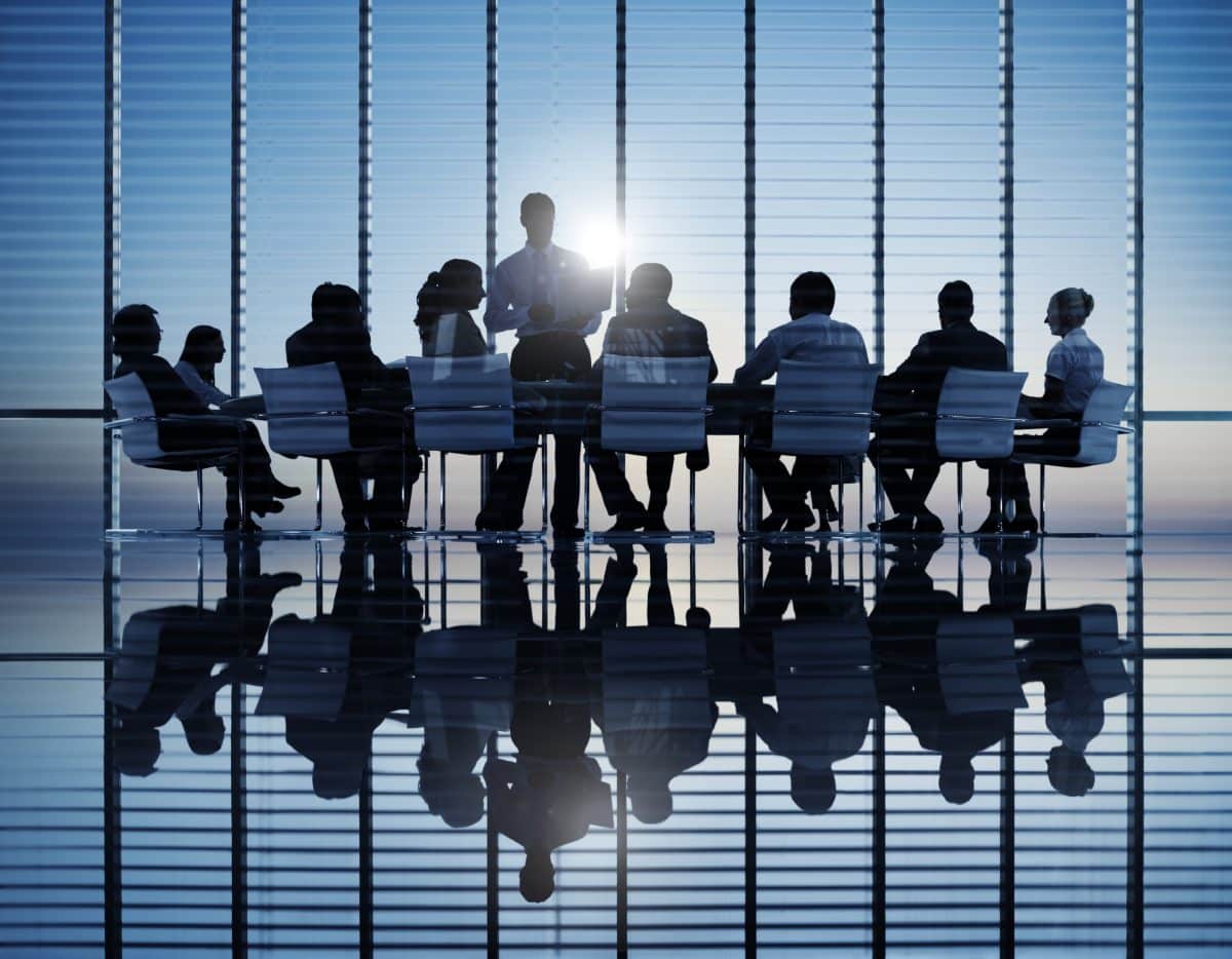 Silhouette d'un groupe de personnes assises autour d'une table de conférence dans un bureau moderne avec de grandes fenêtres et une vue sur la ville, une personne debout et parlant.