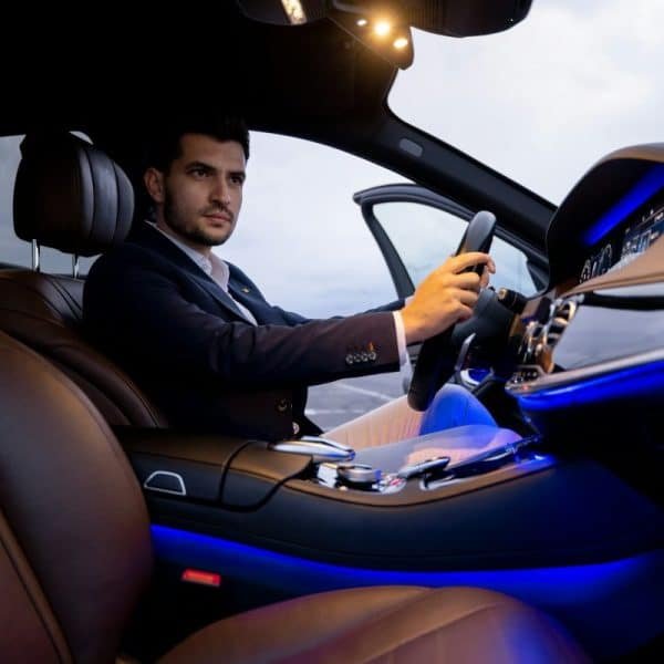 Un homme en costume est assis au siège conducteur d'une voiture de luxe au crépuscule, avec la porte de la voiture ouverte et les lumières intérieures allumées.
