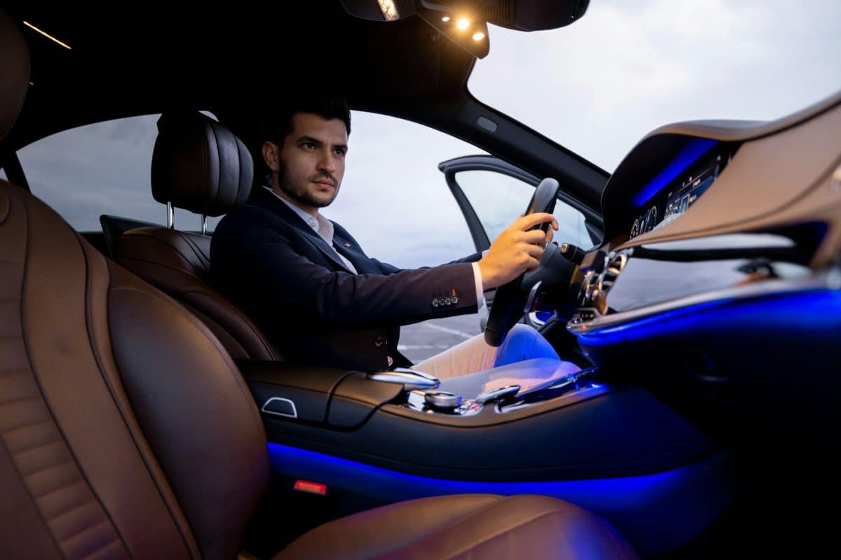 Un homme en costume est assis au siège conducteur d'une voiture de luxe au crépuscule, avec la porte de la voiture ouverte et les lumières intérieures allumées.