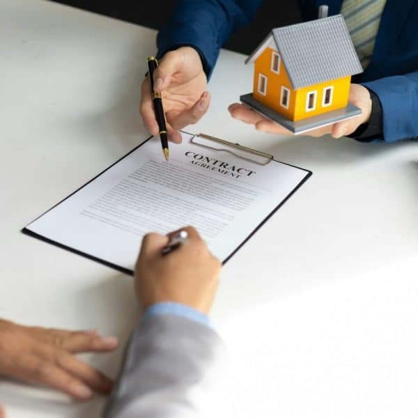 Deux personnes signent un contrat immobilier à une table. L'une d'elles tient une petite maquette de maison tout en offrant un stylo à l'autre personne, qui signe le document.
