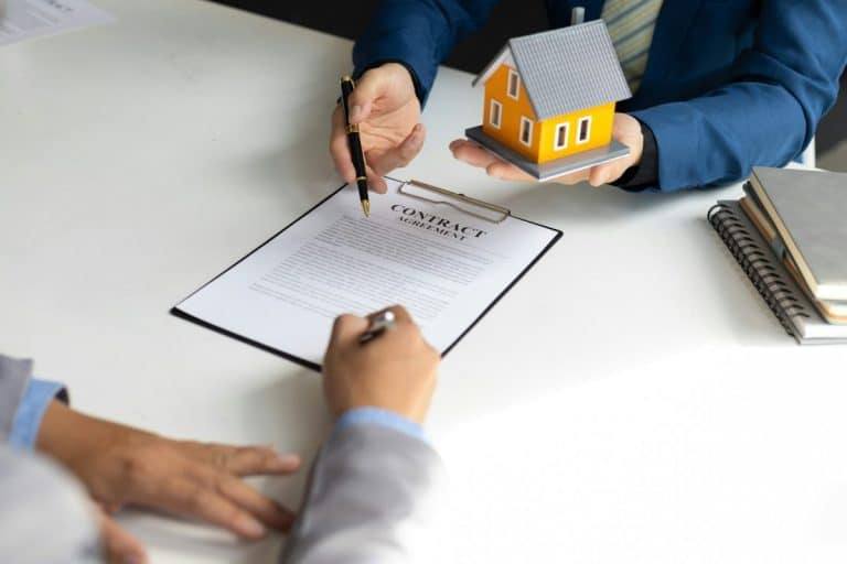Deux personnes signent un contrat immobilier à une table. L'une d'elles tient une petite maquette de maison tout en offrant un stylo à l'autre personne, qui signe le document.