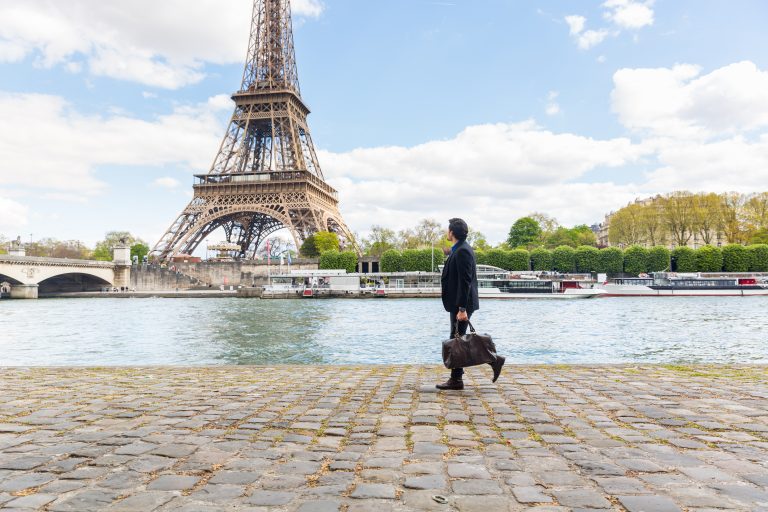 Une personne portant un sac marche le long de la Seine avec la Tour Eiffel en arrière plan par une journée partiellement nuageuse à Paris.