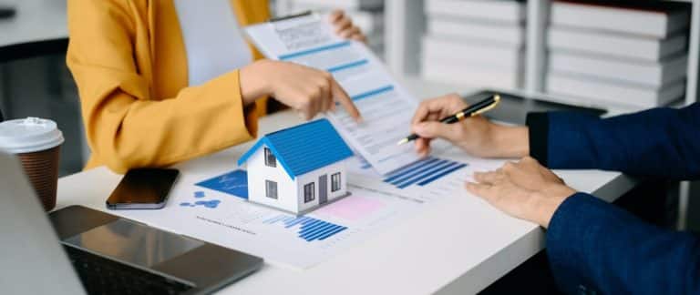 Deux professionnels discutent de documents avec une petite maison modèle sur la table, indiquant une réunion de planification immobilière ou financière.