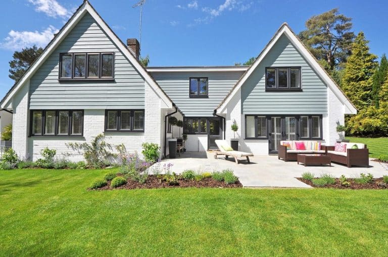 Une maison à deux étages avec un revêtement gris et des accents blancs, dotée d'un patio avec des sièges extérieurs, située sur une pelouse paysagée avec des buissons et des arbres en arrière plan sous un ciel bleu clair.