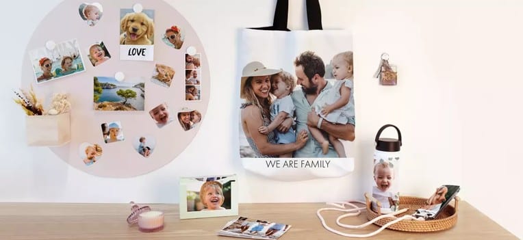 Exposition de cadeaux photo personnalisés, dont un sac fourre tout, une bouteille d'eau et divers tirages, présentant des photos de famille et d'animaux de compagnie sur un mur blanc et une surface en bois.