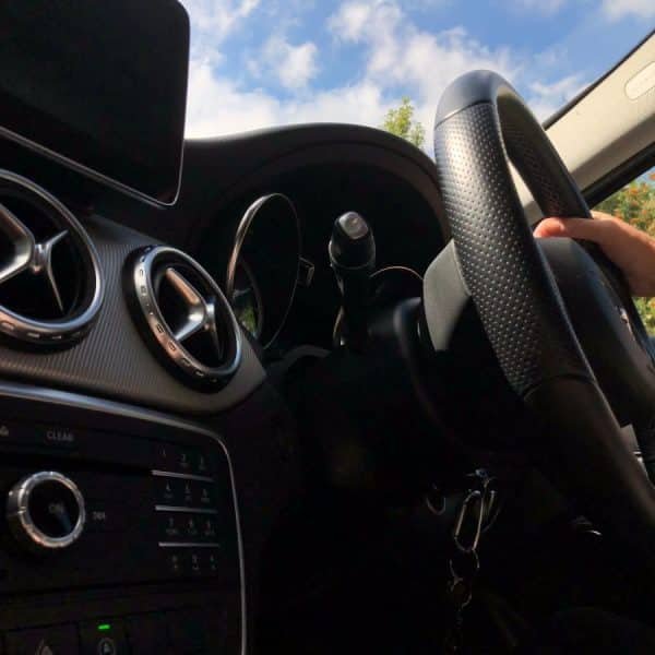 Gros plan de l'intérieur d'une voiture montrant le volant, les commandes du tableau de bord et les bouches d'aération. Une main est posée sur le volant et le ciel est visible à travers le pare brise.