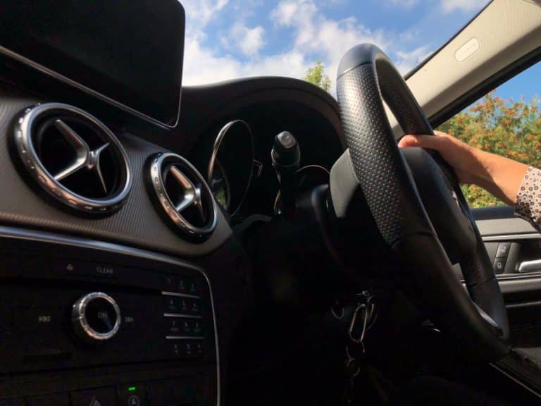 Gros plan de l'intérieur d'une voiture montrant le volant, les commandes du tableau de bord et les bouches d'aération. Une main est posée sur le volant et le ciel est visible à travers le pare brise.