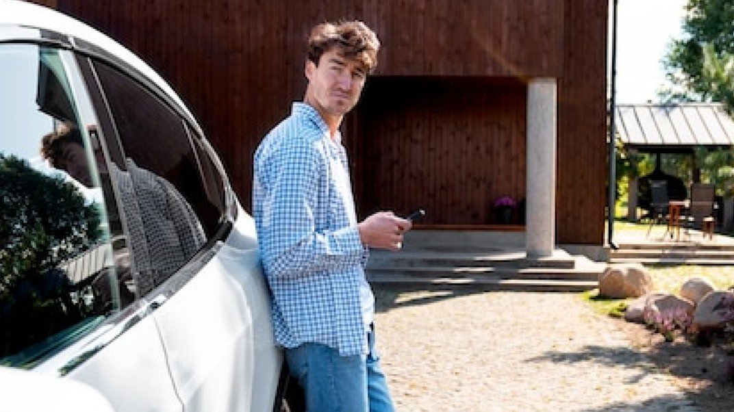 Un homme vêtu d'une chemise bleue à carreaux s'appuie contre une voiture blanche tout en tenant un téléphone, à l'extérieur d'un bâtiment en bois par une journée ensoleillée.