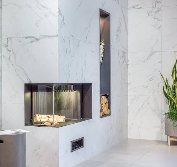 Intérieur moderne avec une cheminée en marbre, du bois de chauffage empilé dans une niche, une grande plante en pot et une poubelle métallique sur un sol carrelé.