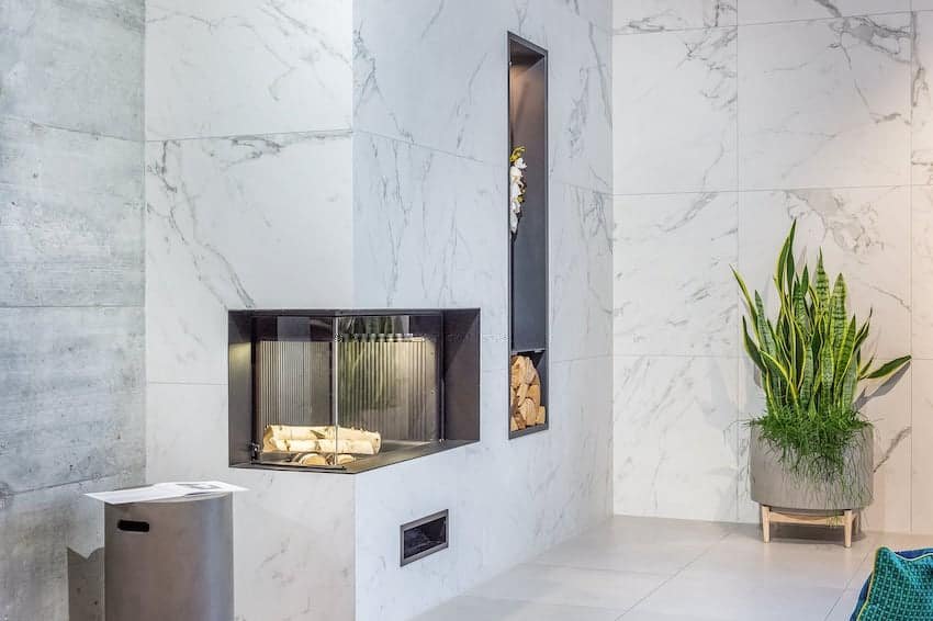Intérieur moderne avec une cheminée en marbre, du bois de chauffage empilé dans une niche, une grande plante en pot et une poubelle métallique sur un sol carrelé.