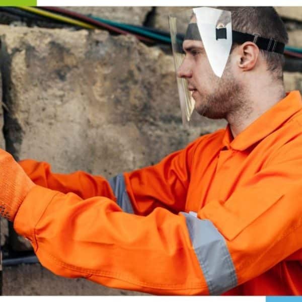Une personne portant un uniforme de sécurité orange et des gants travaille sur un panneau électrique à l’extérieur.
