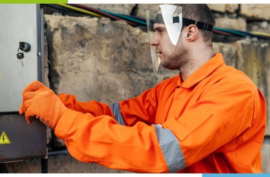 Une personne portant un uniforme de sécurité orange et des gants travaille sur un panneau électrique à l’extérieur.