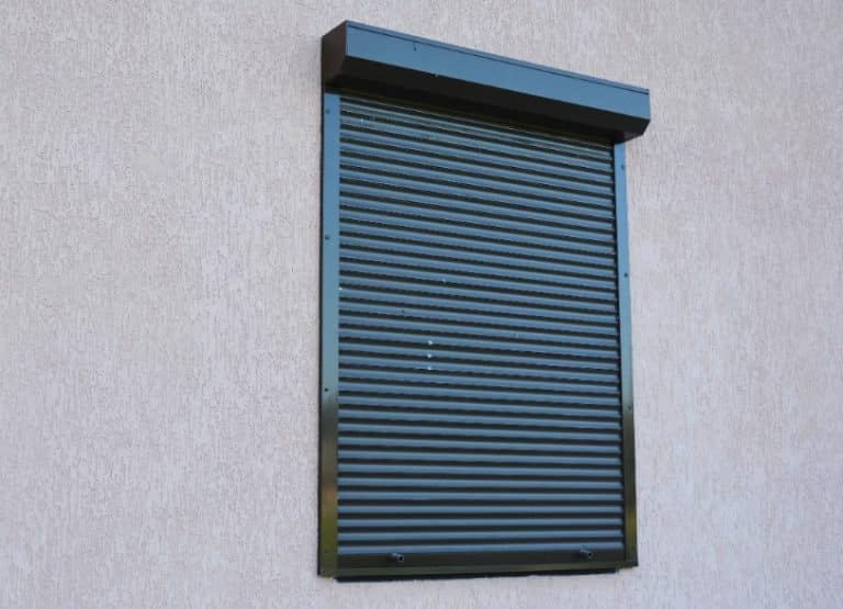 Un volet de sécurité en métal noir fermé est monté au dessus d'une fenêtre sur un mur extérieur de couleur claire.
