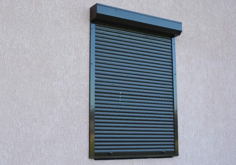 Un volet de sécurité en métal noir fermé est monté au dessus d'une fenêtre sur un mur extérieur de couleur claire.