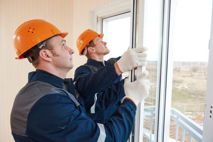 Deux ouvriers du bâtiment portant des casques et des gants orange installent une fenêtre à l'intérieur d'un bâtiment, avec une scène extérieure visible à travers la vitre.