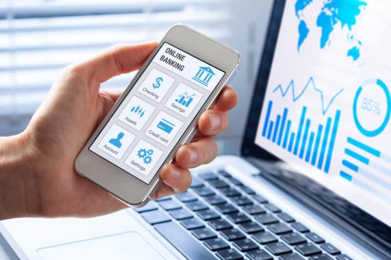 Une personne tenant un smartphone affichant une application bancaire en ligne avec des options telles que les chèques, l'épargne et plus encore, devant un ordinateur portable affichant des graphiques financiers et une statistique de 85 % à l'écran.