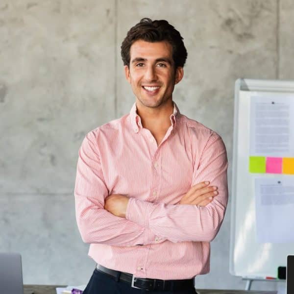 Un homme vêtu d'une chemise rose se tient debout, les bras croisés, souriant dans un bureau avec un tableau blanc et des ordinateurs portables en arrière plan.