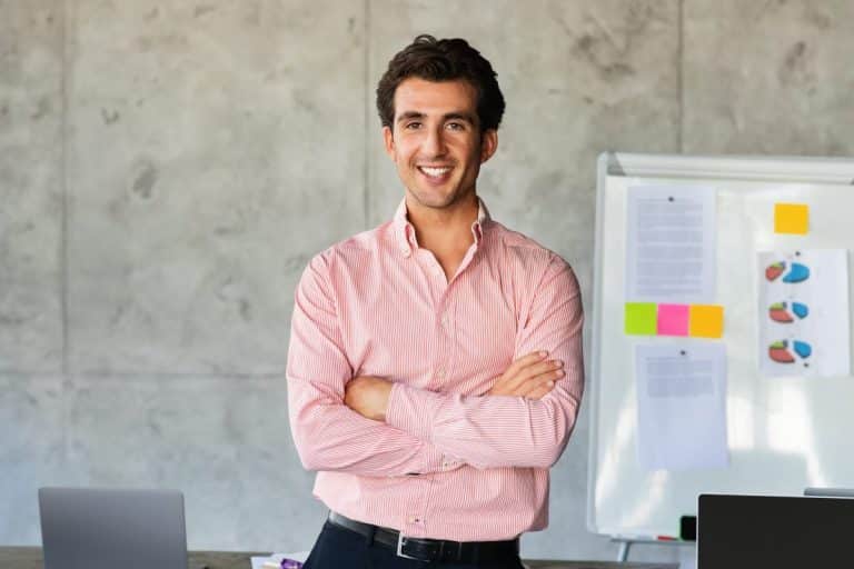 Un homme vêtu d'une chemise rose se tient debout, les bras croisés, souriant dans un bureau avec un tableau blanc et des ordinateurs portables en arrière plan.