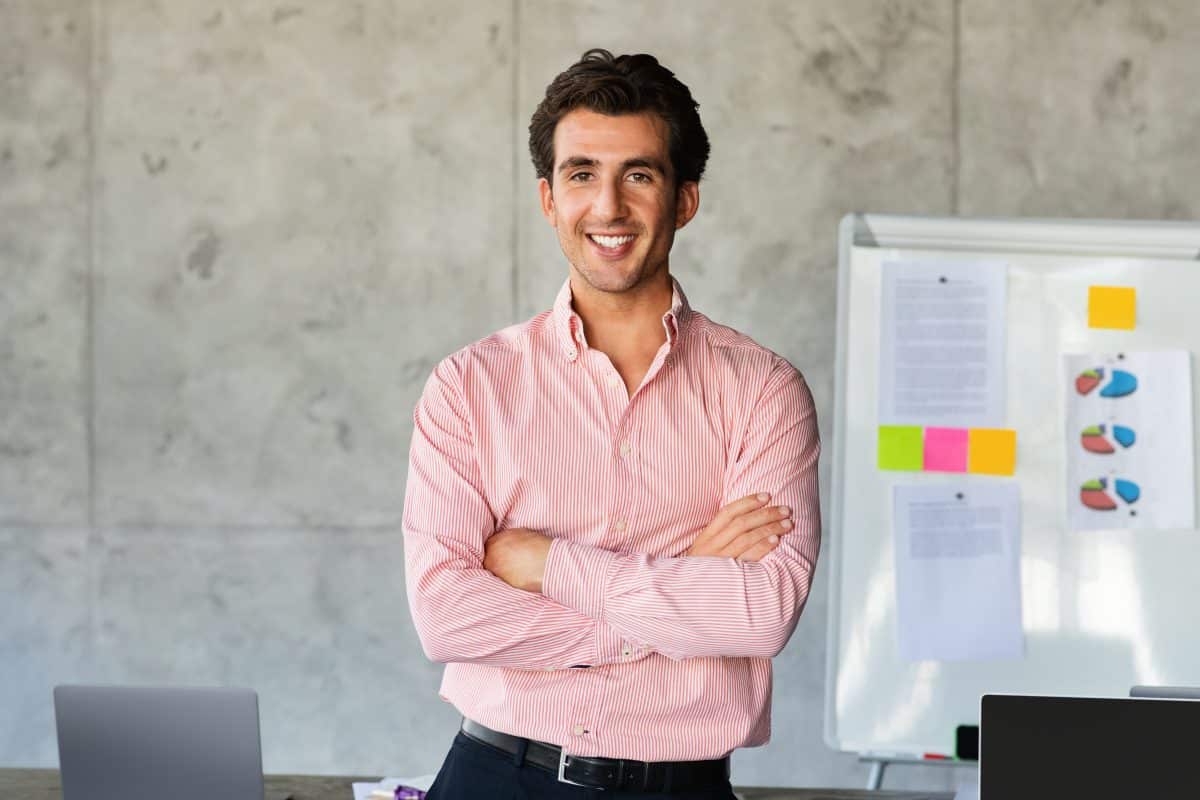 Un homme vêtu d'une chemise rose se tient debout, les bras croisés, souriant dans un bureau avec un tableau blanc et des ordinateurs portables en arrière plan.