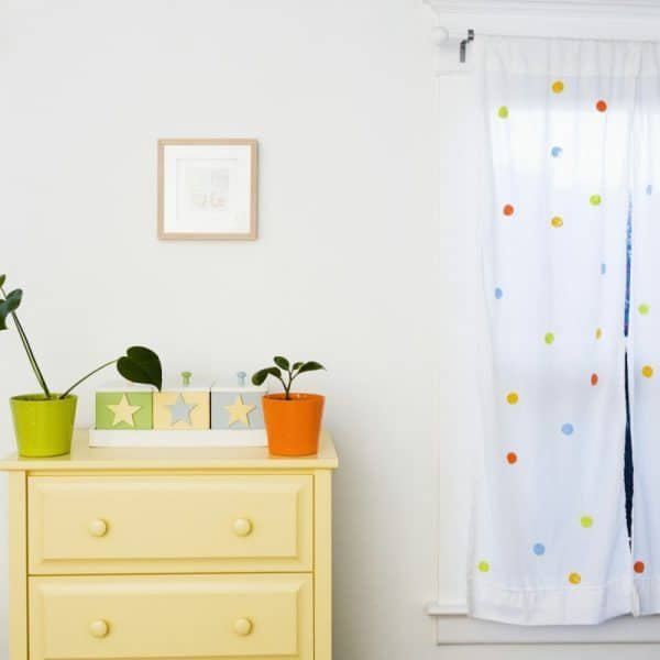 Une commode jaune clair avec des plantes vertes et oranges en pot est placée entre deux fenêtres aux rideaux blancs à pois colorés. Un petit tableau encadré est suspendu au-dessus de la commode.