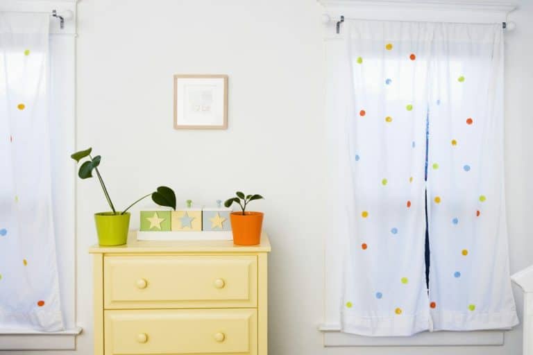 Une commode jaune clair avec des plantes vertes et oranges en pot est placée entre deux fenêtres aux rideaux blancs à pois colorés. Un petit tableau encadré est suspendu au-dessus de la commode.