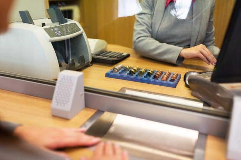 Un caissier de banque est assis derrière une vitre avec un comptoir d'argent, un plateau de pièces et une calculatrice sur le bureau, servant un client.