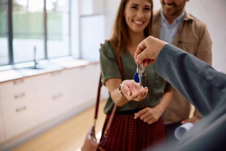 Un couple souriant reçoit les clés de la maison d'une personne, debout dans une cuisine lumineuse.