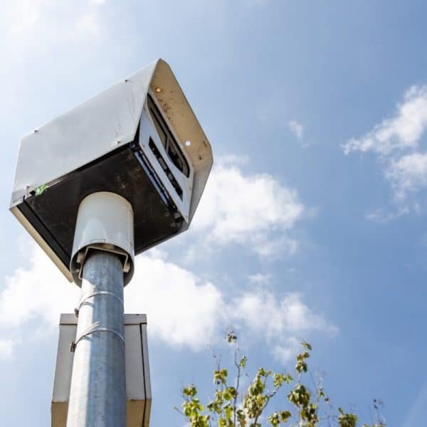Caméra de circulation montée sur un grand poteau contre un ciel bleu clair avec quelques nuages et quelques branches d'arbres visibles au premier plan.