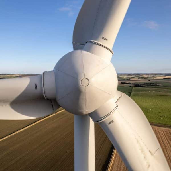 Vue rapprochée des pales et du moyeu du rotor d'une éolienne sur fond de vastes champs agricoles sous un ciel bleu clair.