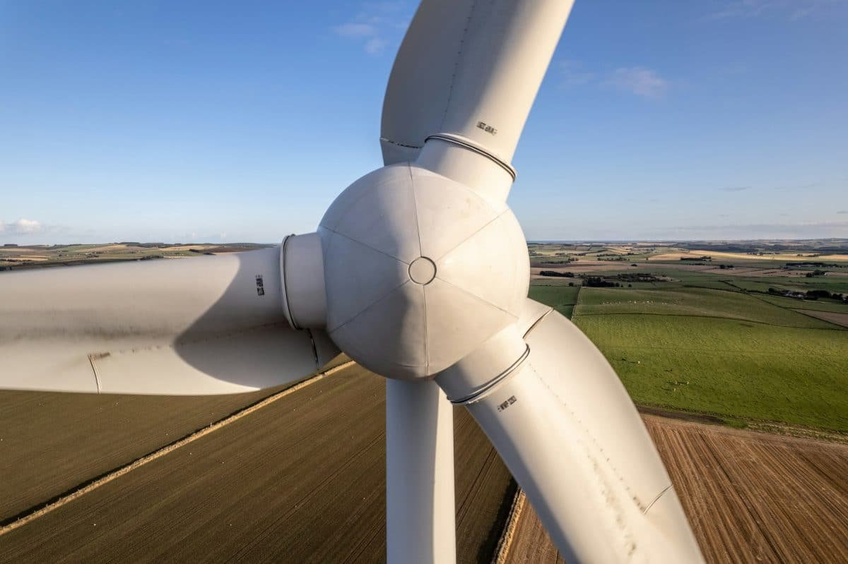Vue rapprochée des pales et du moyeu du rotor d'une éolienne sur fond de vastes champs agricoles sous un ciel bleu clair.