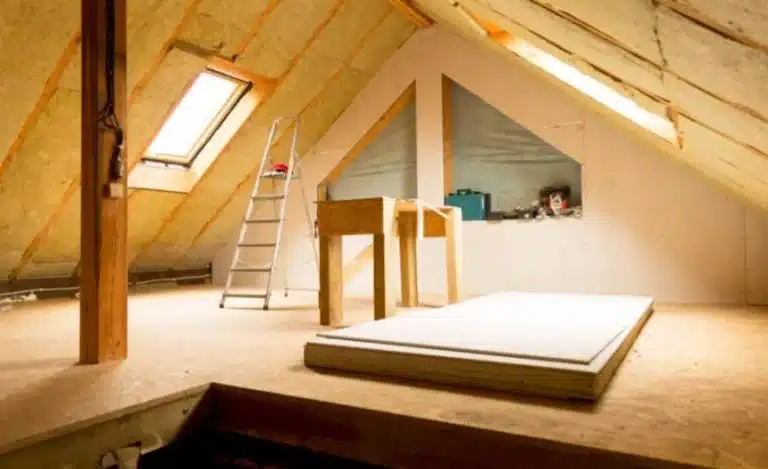 Grenier non aménagé avec isolation apparente, puits de lumière, échelle, établi et matériaux de construction au sol.