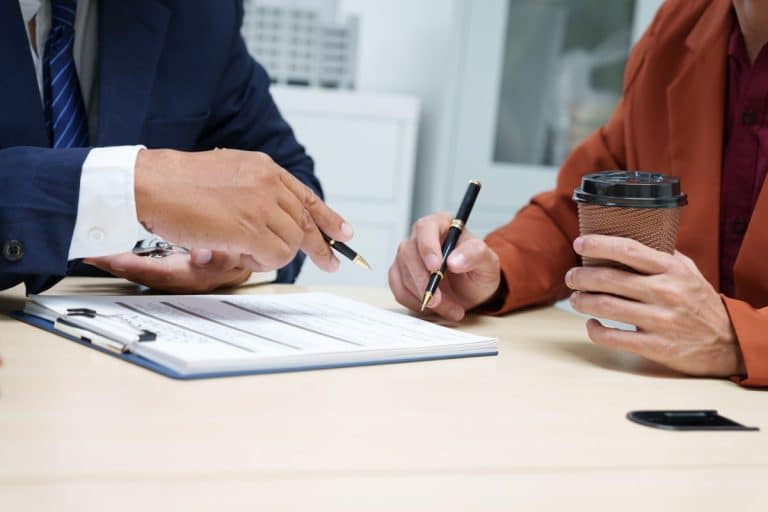 Deux personnes en tenue de travail sont assises à un bureau et discutent de documents sur un presse papiers ; l'une tient un stylo, l'autre une tasse à café.
