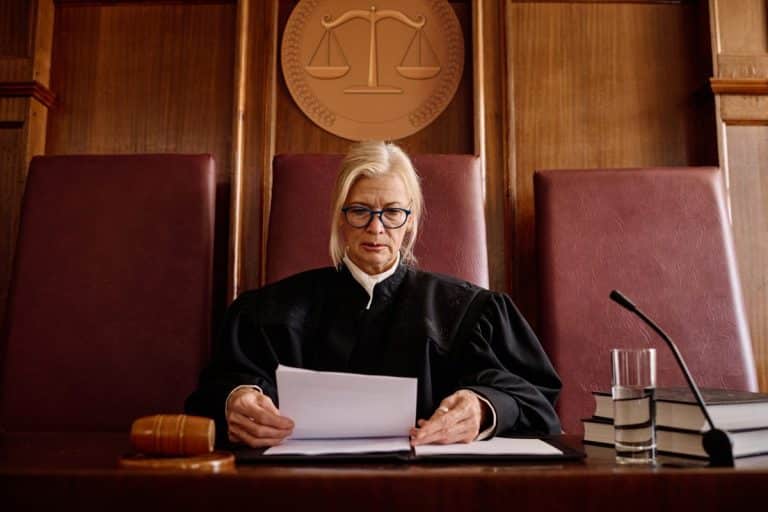 Un juge en robe noire est assis à un bureau et lit des documents. Un marteau, un verre d'eau et des livres se trouvent à proximité, tandis qu'un emblème de la balance de la justice est accroché au mur derrière lui.